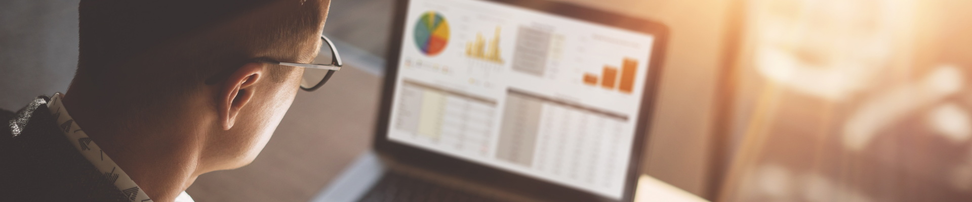 Young finance market analyst in eyeglasses working at sunny office on laptop while sitting at wooden table.Businessman analyze document in his hands.Graphs and diagramm on notebook screen.Blurred