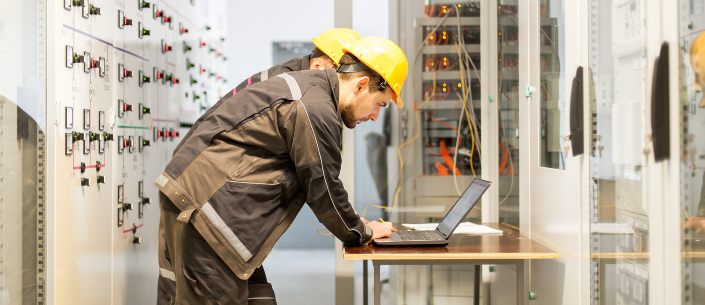 Two maintenance engineers inspect relay protection system with laptop computer. Bay control unit. Medium voltage switchgear